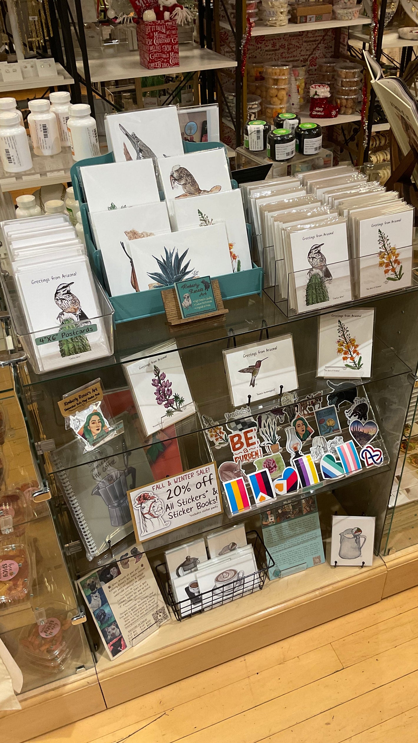 Collection of Kimberly Renee's Art greeting cards, prints, and stickers on a glass shelf with a wooden floor background. Arizona and coffee themes.