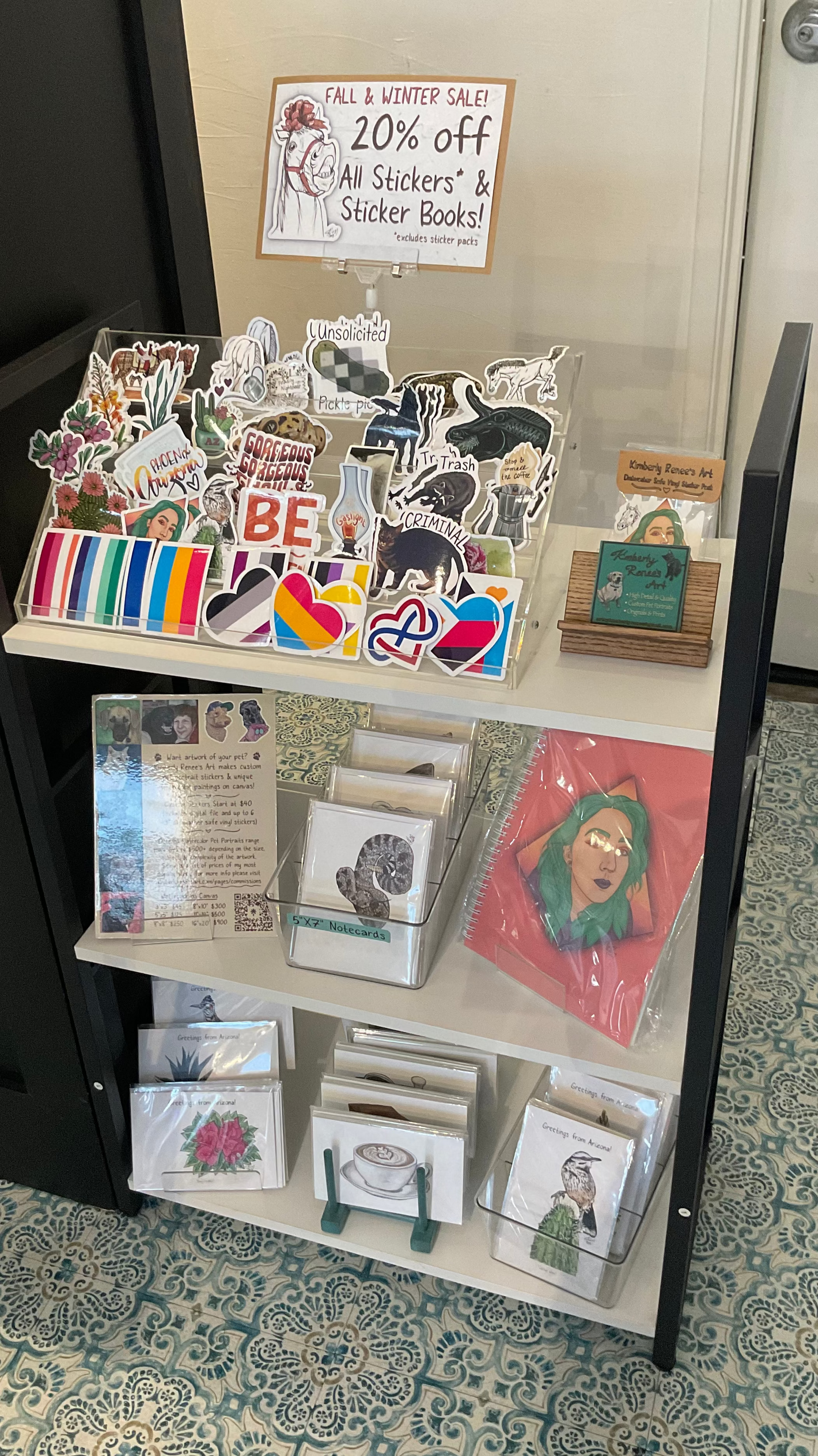 Display of Kimberly Renee's Art stickers and greeting cards with a sale sign in the background. Arizona, coffee, and animal themes.